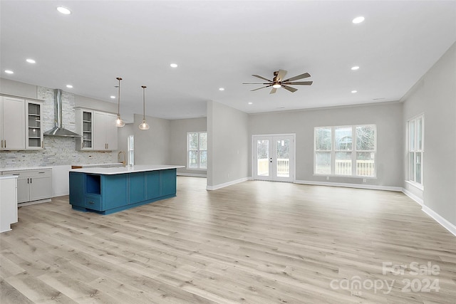 kitchen with wall chimney exhaust hood, backsplash, an island with sink, pendant lighting, and light wood-type flooring