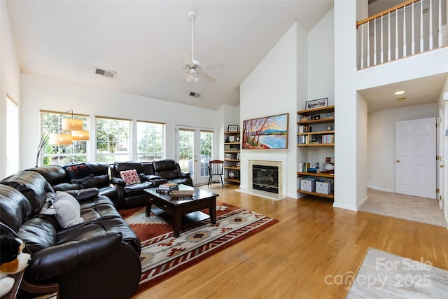living room with visible vents, ceiling fan, light wood finished floors, high vaulted ceiling, and a glass covered fireplace