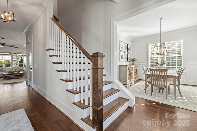 staircase with crown molding, a decorative wall, hardwood / wood-style floors, baseboards, and ceiling fan with notable chandelier