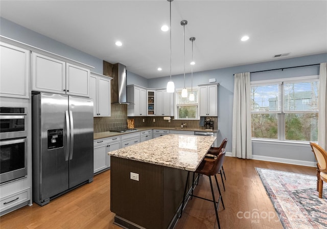 kitchen with appliances with stainless steel finishes, wall chimney exhaust hood, light hardwood / wood-style flooring, a center island, and hanging light fixtures