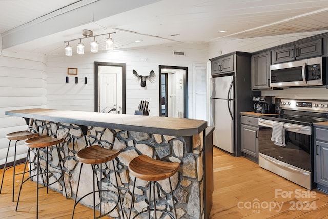 kitchen featuring stainless steel appliances, vaulted ceiling, wooden walls, light hardwood / wood-style flooring, and a breakfast bar area