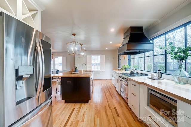 kitchen featuring a kitchen island with sink, white cabinets, butcher block countertops, custom range hood, and stainless steel appliances