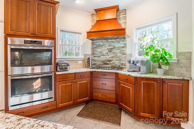 kitchen with light stone counters, backsplash, crown molding, custom exhaust hood, and appliances with stainless steel finishes