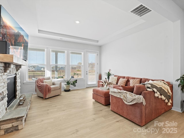 living area featuring light wood-style floors, visible vents, a tray ceiling, and a stone fireplace