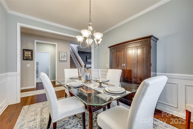 dining space with a wainscoted wall, dark wood-type flooring, an inviting chandelier, stairs, and crown molding