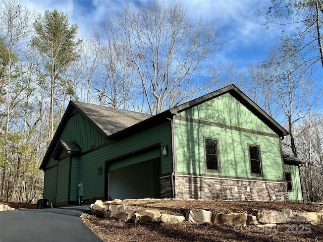 view of side of property featuring stone siding, driveway, an attached garage, and roof with shingles