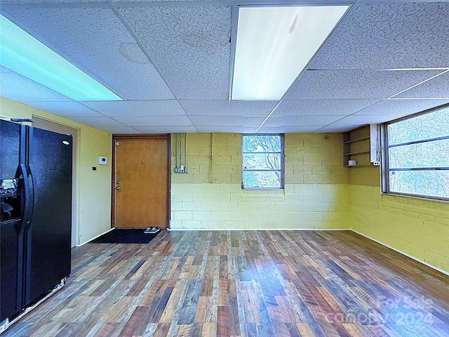 basement featuring black fridge with ice dispenser, a drop ceiling, plenty of natural light, and hardwood / wood-style flooring