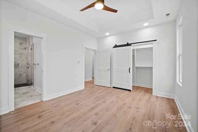 unfurnished bedroom featuring ceiling fan, a raised ceiling, a barn door, ensuite bathroom, and light wood-type flooring