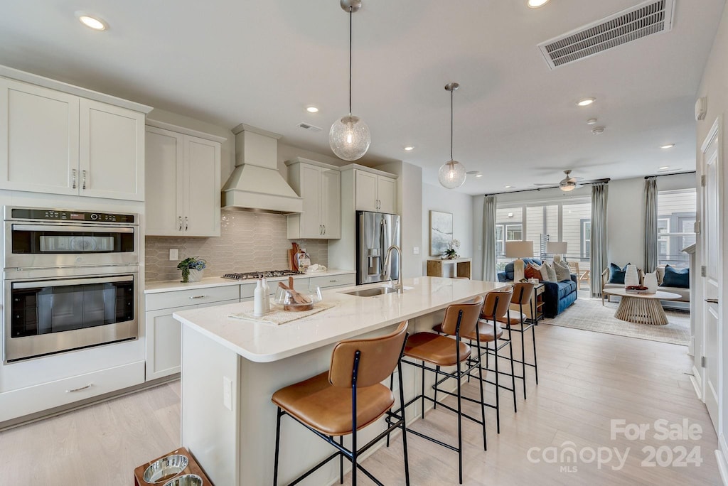 kitchen featuring custom exhaust hood, light hardwood / wood-style flooring, an island with sink, appliances with stainless steel finishes, and decorative light fixtures