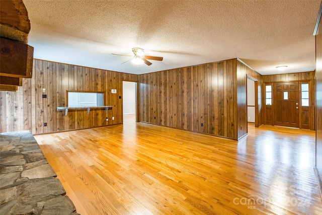 unfurnished living room with ceiling fan, light wood-type flooring, a textured ceiling, and wooden walls