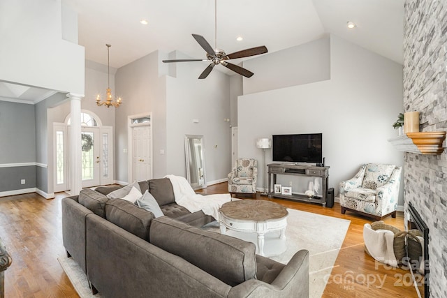 living room featuring light wood-type flooring, decorative columns, ceiling fan with notable chandelier, high vaulted ceiling, and a stone fireplace