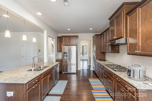 kitchen with light stone countertops, appliances with stainless steel finishes, a kitchen island with sink, dark wood-type flooring, and sink