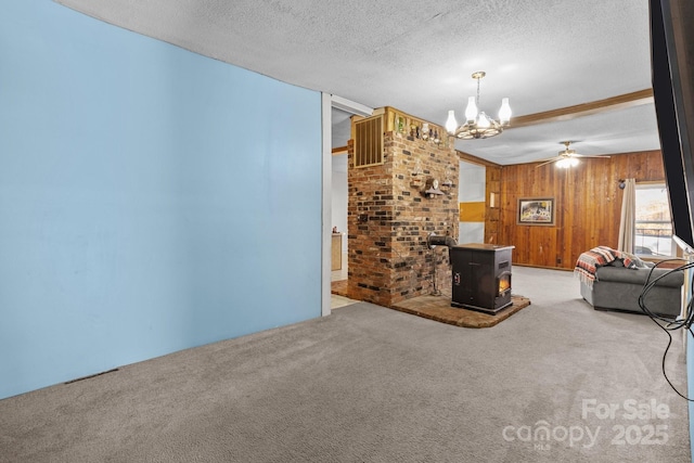 unfurnished living room with ceiling fan with notable chandelier, a textured ceiling, wooden walls, carpet floors, and a wood stove