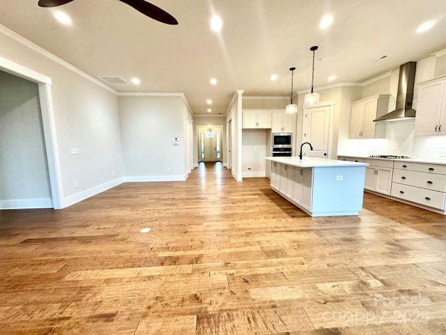 kitchen featuring wall chimney range hood, tasteful backsplash, an island with sink, stainless steel gas stovetop, and white cabinets