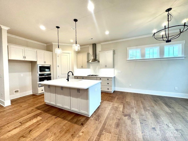 kitchen featuring white cabinetry, wall chimney range hood, decorative light fixtures, a center island with sink, and appliances with stainless steel finishes