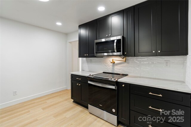 kitchen featuring appliances with stainless steel finishes, dark cabinets, light wood finished floors, decorative backsplash, and light stone counters