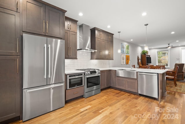 kitchen featuring wall chimney exhaust hood, light wood-type flooring, appliances with stainless steel finishes, decorative light fixtures, and kitchen peninsula