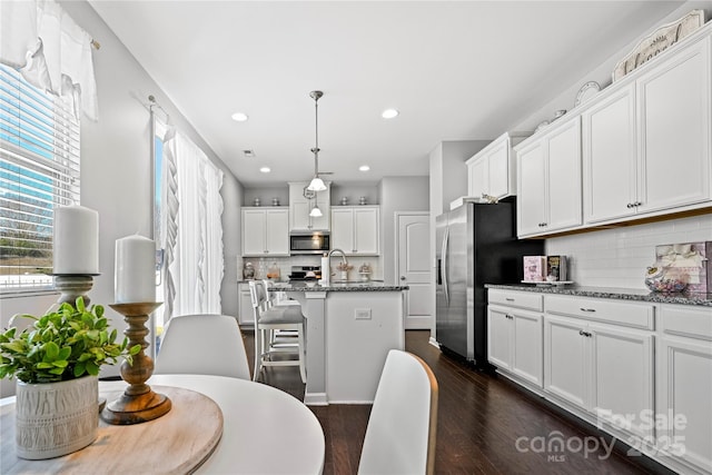 kitchen with stainless steel appliances, white cabinetry, a center island with sink, and dark stone countertops