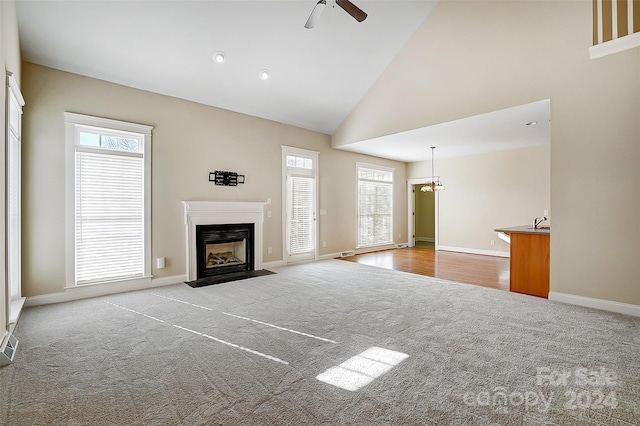 unfurnished living room featuring light carpet, ceiling fan with notable chandelier, high vaulted ceiling, and a healthy amount of sunlight