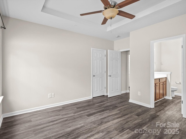 unfurnished bedroom featuring ensuite bathroom, ceiling fan, a tray ceiling, and dark hardwood / wood-style flooring