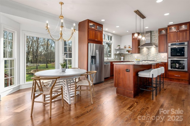 kitchen featuring a kitchen island, glass insert cabinets, decorative light fixtures, stainless steel appliances, and wall chimney range hood