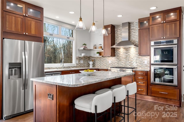 kitchen with light stone countertops, wall chimney exhaust hood, glass insert cabinets, and stainless steel appliances