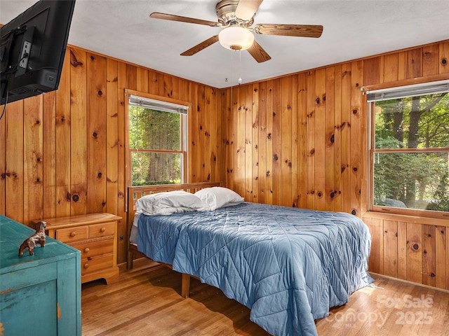 bedroom with ceiling fan, light hardwood / wood-style floors, multiple windows, and wood walls