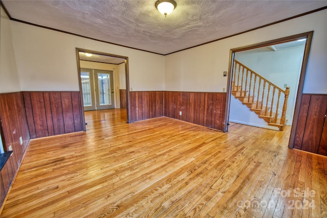 empty room with french doors, light wood-type flooring, ornamental molding, a textured ceiling, and wood walls