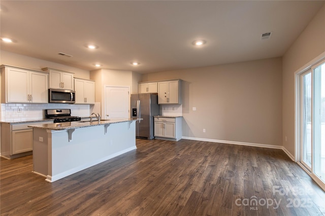 kitchen with appliances with stainless steel finishes, a breakfast bar, dark hardwood / wood-style flooring, light stone counters, and a center island with sink