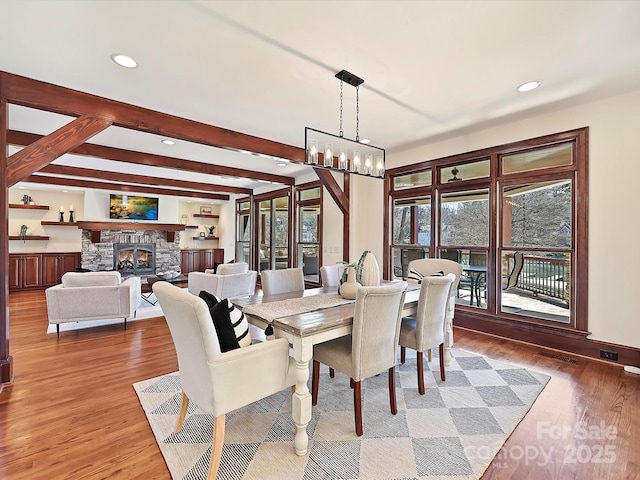 dining space featuring beam ceiling, a stone fireplace, hardwood / wood-style floors, and an inviting chandelier