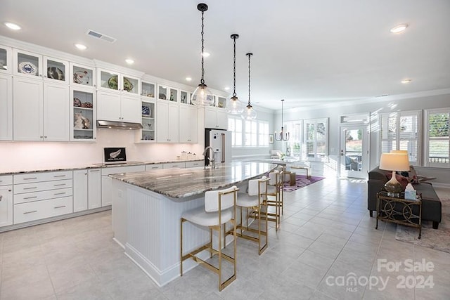 kitchen with white refrigerator with ice dispenser, a kitchen island with sink, hanging light fixtures, ornamental molding, and light stone counters