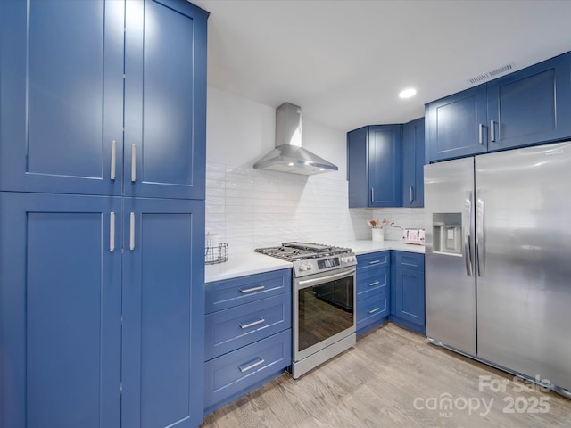 kitchen with stainless steel appliances, tasteful backsplash, blue cabinets, wall chimney exhaust hood, and light wood-type flooring