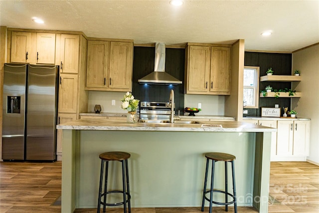 kitchen with a kitchen island with sink, wall chimney range hood, sink, stainless steel fridge, and a breakfast bar area