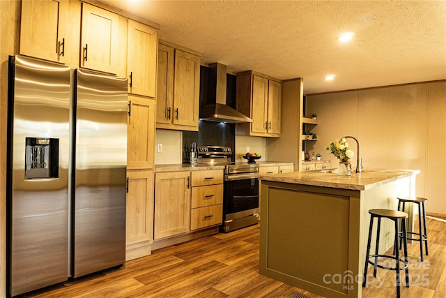 kitchen with wall chimney range hood, sink, an island with sink, light hardwood / wood-style floors, and stainless steel appliances