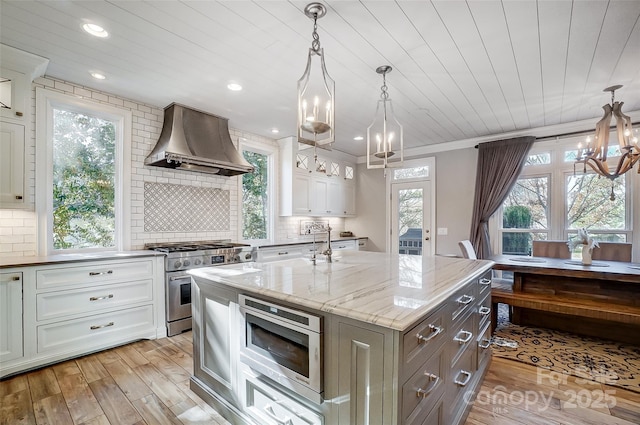 kitchen featuring hanging light fixtures, appliances with stainless steel finishes, an island with sink, light stone countertops, and wall chimney range hood