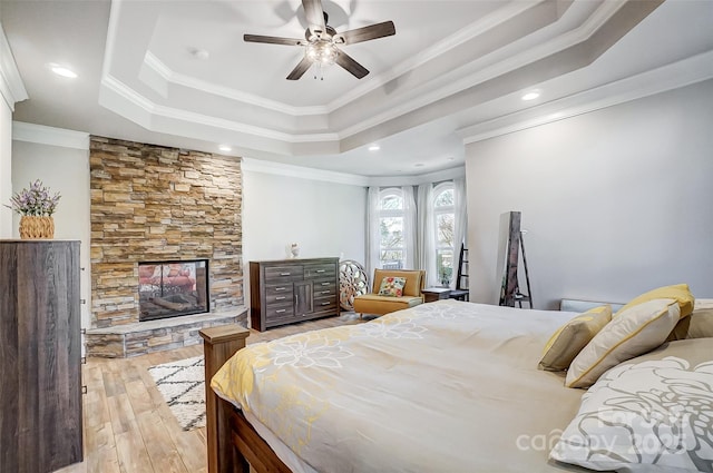 bedroom with a stone fireplace, ceiling fan, a raised ceiling, crown molding, and light wood-type flooring