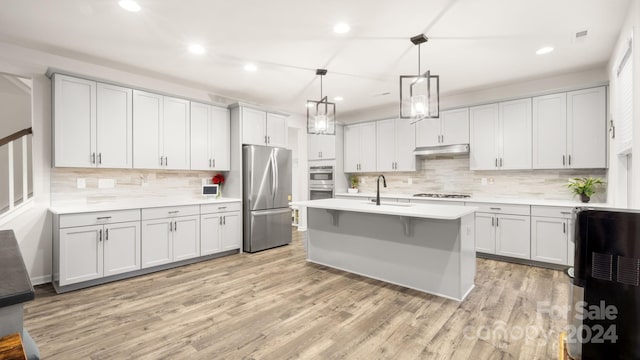 kitchen featuring light hardwood / wood-style flooring, backsplash, decorative light fixtures, a center island with sink, and appliances with stainless steel finishes