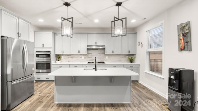 kitchen with hanging light fixtures, stainless steel appliances, a center island with sink, and light hardwood / wood-style floors