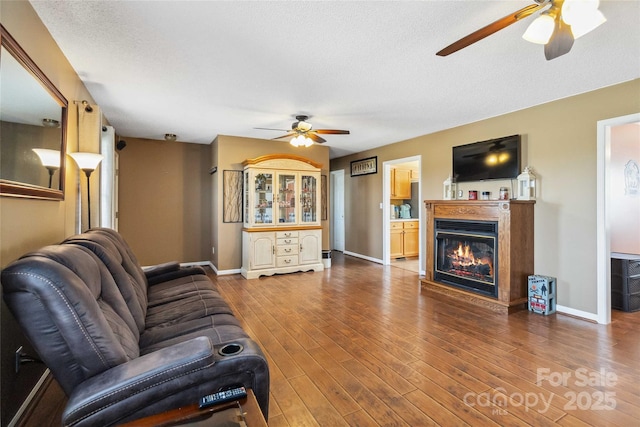 living area featuring a ceiling fan, a glass covered fireplace, wood finished floors, and baseboards