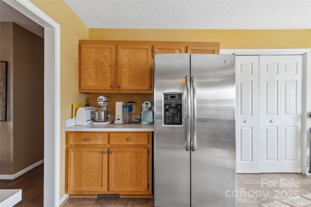 kitchen with stainless steel refrigerator with ice dispenser, light countertops, brown cabinets, a textured ceiling, and baseboards