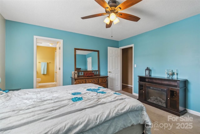 bedroom with baseboards, ensuite bathroom, carpet, a glass covered fireplace, and a ceiling fan