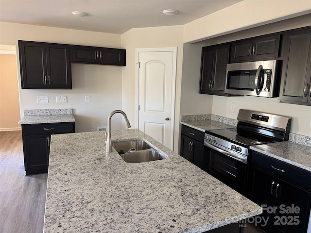 kitchen with sink, a center island with sink, appliances with stainless steel finishes, light stone countertops, and light hardwood / wood-style floors