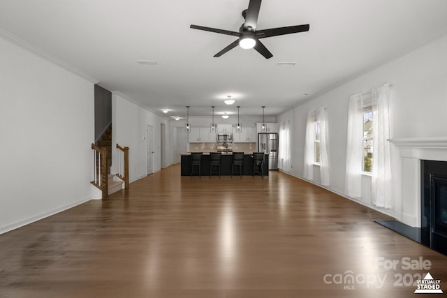 unfurnished living room featuring ceiling fan, stairway, dark wood-style floors, a glass covered fireplace, and crown molding