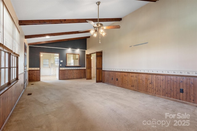 unfurnished living room with ceiling fan, vaulted ceiling with beams, wooden walls, and light colored carpet