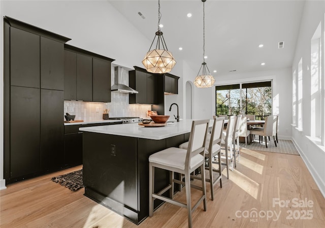 kitchen featuring wall chimney exhaust hood, hanging light fixtures, tasteful backsplash, light hardwood / wood-style floors, and a center island with sink