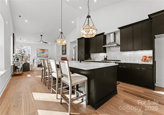 kitchen with tasteful backsplash, sink, wall chimney range hood, pendant lighting, and a center island with sink