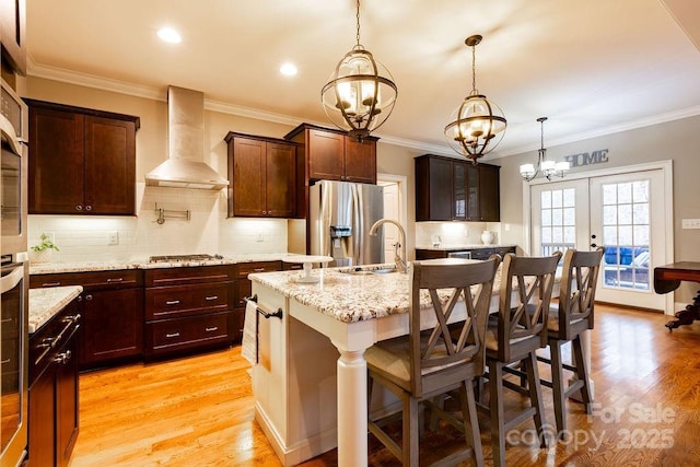 kitchen featuring french doors, light wood-type flooring, wall chimney exhaust hood, a breakfast bar area, and an island with sink