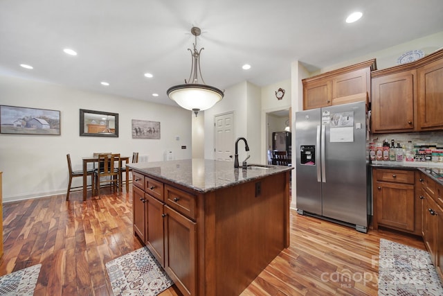 kitchen with pendant lighting, a kitchen island with sink, sink, light wood-type flooring, and stainless steel fridge with ice dispenser