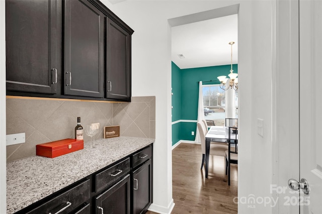 kitchen featuring wood-type flooring, a chandelier, dark brown cabinets, light stone countertops, and backsplash