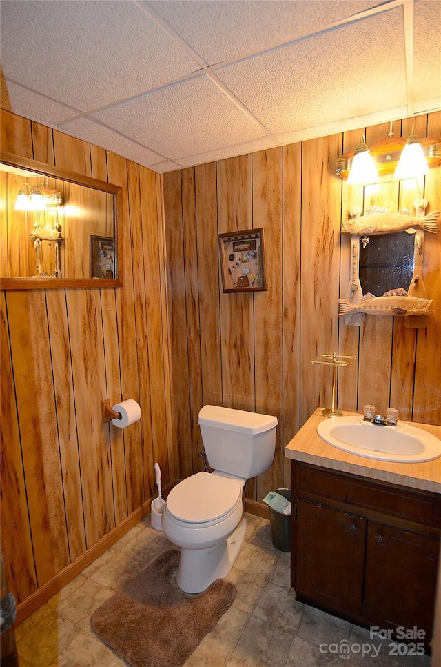 bathroom with wood walls, a drop ceiling, vanity, and toilet